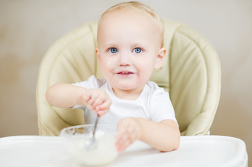 Smiling girl is picking up porridge with a spoon while sitting in a baby seat for feeding