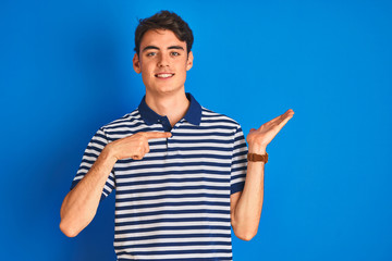 Teenager boy wearing casual t-shirt standing over blue isolated background amazed and smiling to the camera while presenting with hand and pointing with finger.