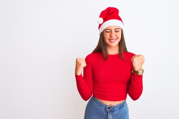 Young beautiful girl wearing Christmas Santa hat standing over isolated white background very happy and excited doing winner gesture with arms raised, smiling and screaming for success