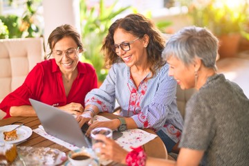 Meeting of middle age women having lunch and drinking coffee. Mature friends smiling happy using laptop at home on a sunny day