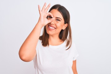 Portrait of beautiful and young brunette woman standing over isolated white background doing ok gesture with hand smiling, eye looking through fingers with happy face.