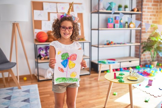 Beautiful toddler wearing glasses and unicorn diadem standing holding draw smiling at kindergarten