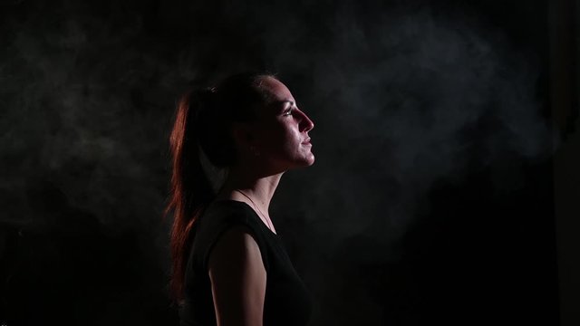A sad brunette is in a cloud of smoke. Portrait of a woman smoking a vape behind her, soaring. Black background. Passive smoking. Bad habits.