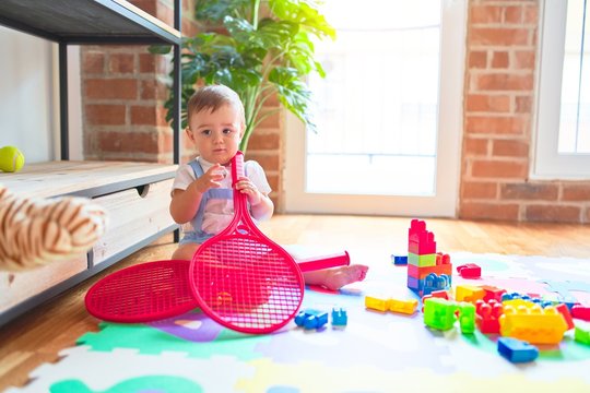 Beautiful toddler sitting on puzzle carpet holding tennis racket at kindergarten