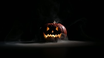Halloween, orange pumpkin with a scary luminous face on a dark background. Thick gray smoke comes out and spreads across the black table. A close-up of a flashlight on the eve of all the saints