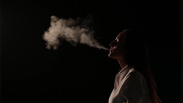 Pensive brunette in a white shirt smokes a hookah in the dark. Portrait of a woman smoking a vape, hovering. Black background. Electronic Cigarette. Bad habit.