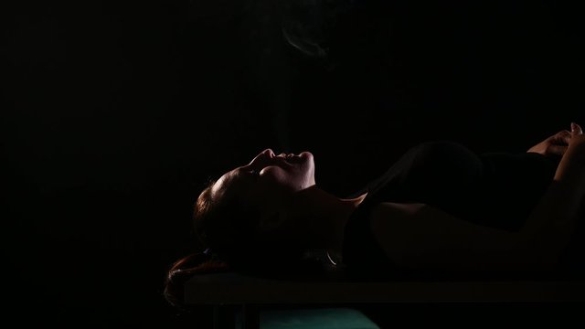 The brunette smokes an electronic cigarette on a black background, releases thick smoke from her mouth. Portrait of a woman smoking a vape, hovering.