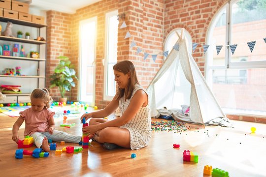 Beautiful teacher and blond toddler girl building tower using plastic blocks at kindergarten