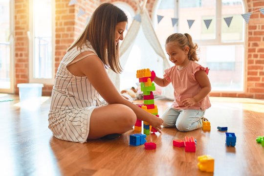 Beautiful teacher and blond toddler girl building tower using plastic blocks at kindergarten