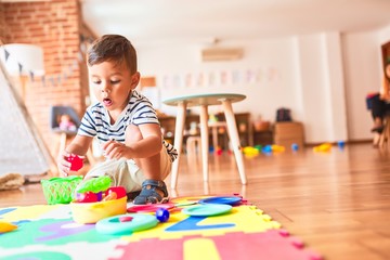 Beautiful toddler boy sitting on puzzle playing meals with plastic plates, fruits and vegetables at kindergarten