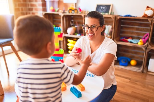 Beautiful teacher and toddler boy playing with construction blocks bulding tower at kindergarten
