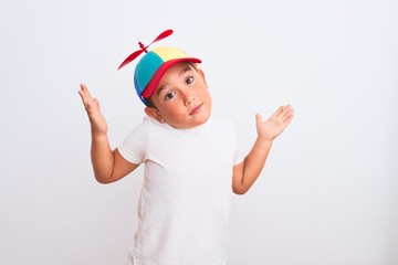 Beautiful kid boy wearing fanny colorful cap with propeller over isolated white background clueless and confused expression with arms and hands raised. Doubt concept. © Krakenimages.com