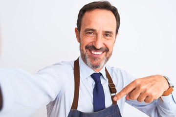 Middle age shopkeeper man wearing apron make selfie over isolated white background very happy pointing with hand and finger