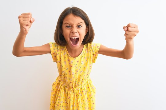 Young beautiful child girl wearing yellow floral dress standing over isolated white background angry and mad raising fists frustrated and furious while shouting with anger. Rage