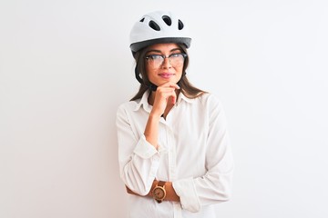 Beautiful businesswoman wearing glasses and bike helmet over isolated white background looking confident at the camera smiling with crossed arms and hand raised on chin. Thinking positive.