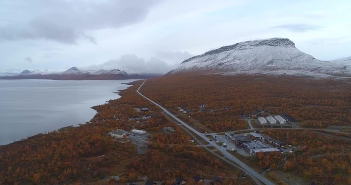 Kilpisjarvi town, Aerial, tracking, drone shot, overlooking the lake, the e8 road, the village and first snow on Saana tunturi mountain, on a cold, autumn morning, in Enontekio, Finland