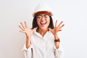 Young beautiful architect woman wearing helmet and glasses over isolated white background celebrating mad and crazy for success with arms raised and closed eyes screaming excited. Winner concept