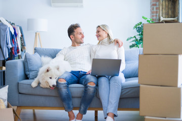 Fototapeta premium Young beautiful couple with dog sitting on the sofa using laptop at new home around cardboard boxes