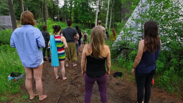 Diverse people enjoy spiritual gathering A mixed group of individuals are seen standing barefoot in a forest clearing, practicing traditional yoga and native spirituality exercises by a tipi tent.