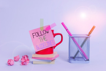 Word writing text Follow The Guidelines. Business photo showcasing Manual of Style Follow a Specified Rule Accordingly Cup pens holder note banners stacked pads paper balls pastel background