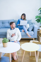 Young beautiful couple sitting on the sofa drinking cup of coffee using laptop at new home around cardboard boxes