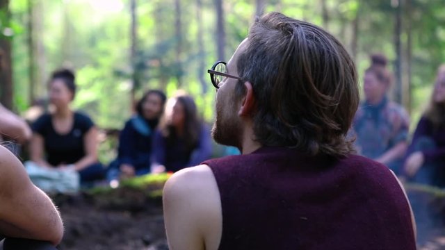 Diverse people enjoy spiritual gathering Slow motion footage of a slim caucasian man, wearing glasses and sleeveless top, sitting by a smoldering campfire during a shamanic retreat in nature.