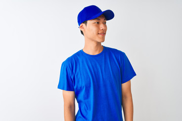 Chinese deliveryman wearing blue t-shirt and cap standing over isolated white background looking away to side with smile on face, natural expression. Laughing confident.