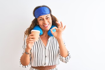 Tourist woman wearing neckpillow sleep mask drinking coffee over isolated white background doing ok sign with fingers, excellent symbol