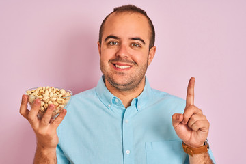 Young man holding bowl with pistachios standing over isolated pink background surprised with an idea or question pointing finger with happy face, number one