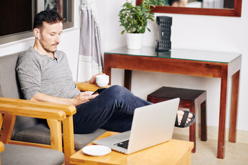 Young man enjoying cup of coffee and scrolling social media on smartphone after working on laptop at home