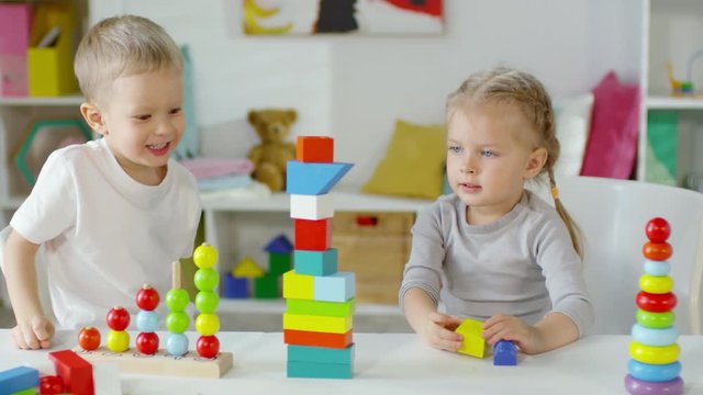 Little brother and sister of preschool age making block tower together and smiling while playing with educational toy at table in playroom
