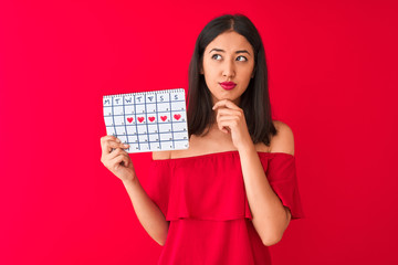 Young beautiful chinese woman holding period calendar standing over isolated red background serious face thinking about question, very confused idea