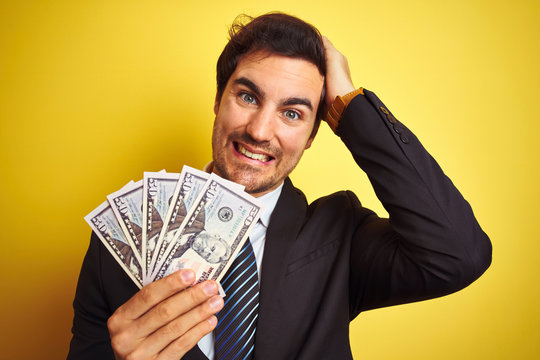 Young handsome businessman wearing suit holding dollars over isolated yellow background stressed with hand on head, shocked with shame and surprise face, angry and frustrated. Fear
