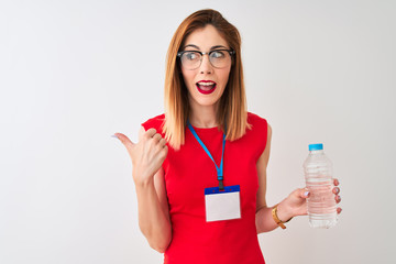 Redhead businesswoman wearing id card drinking water over isolated white background pointing and showing with thumb up to the side with happy face smiling