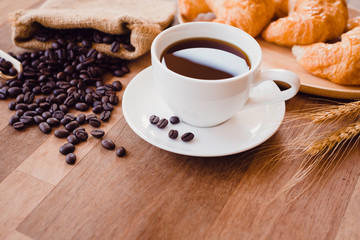 Top view white hot coffee cup and beans on wooden table with croissant bread background.Black coffee menu for breakfast in the coffee shop.Selective focus.