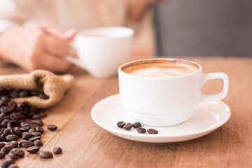 Hot coffee in the white cup and coffee beans in a sack on wooden table background with blurry woman holding coffee cup.Cafe drinking menu latte art coffee at the restaurant with copy space.