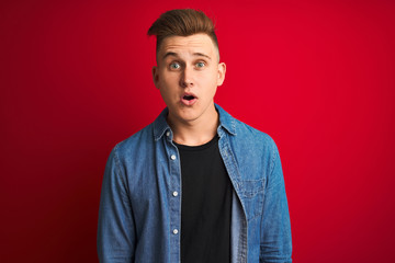 Young handsome man wearing denim shirt standing over isolated red background afraid and shocked with surprise expression, fear and excited face.