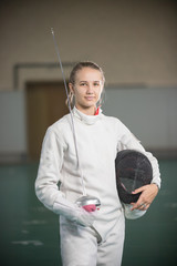 A young smiling woman fencer standing in the gym holding sword and helmet
