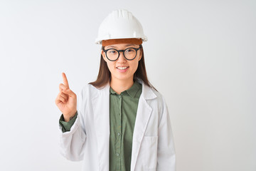 Young chinese engineer woman wearing coat helmet glasses over isolated white background showing and pointing up with finger number one while smiling confident and happy.