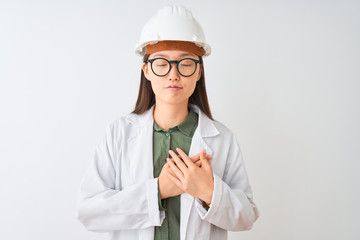 Young chinese engineer woman wearing coat helmet glasses over isolated white background smiling with hands on chest with closed eyes and grateful gesture on face. Health concept.