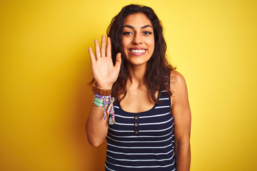 Young beautiful woman wearing striped t-shirt standing over isolated yellow background Waiving saying hello happy and smiling, friendly welcome gesture