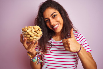 Young beautiful woman holding bowl with peanuts over isolated pink background happy with big smile doing ok sign, thumb up with fingers, excellent sign