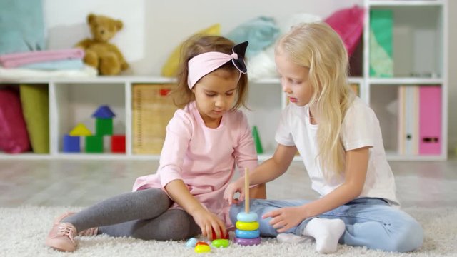 Two pretty little girls of preschool age sitting on the floor in playroom and building colorful toy pyramid together