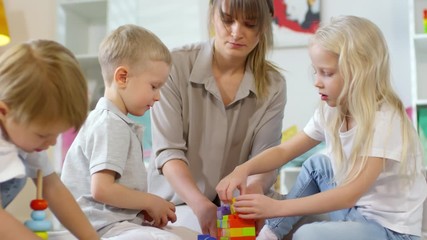 Mother building toy tower with colorful bricks and speaking to little daughter and son while playing with kids on the floor at home