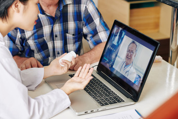 Fototapeta na wymiar Cropped image of medical nurse and senior patient sitting at table with opened laptop and consulting with general practitioner about pills and painkillers