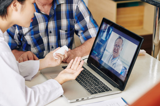 Cropped image of medical nurse and senior patient sitting at table with opened laptop and consulting with general practitioner about pills and painkillers