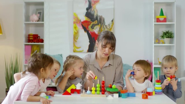 Female teacher sitting at table in kindergarten with group of preschool children, showing them colorful beads and explaining how to play with educational toy