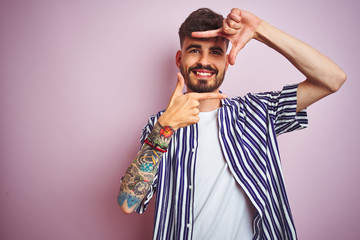 Young man with tattoo wearing striped shirt standing over isolated pink background smiling making frame with hands and fingers with happy face. Creativity and photography concept.