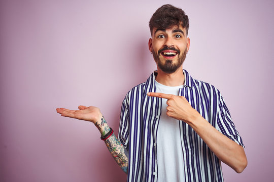 Young man with tattoo wearing striped shirt standing over isolated pink background amazed and smiling to the camera while presenting with hand and pointing with finger.