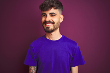 Young man with tattoo wearing t-shirt standing over isolated purple background looking away to side with smile on face, natural expression. Laughing confident.